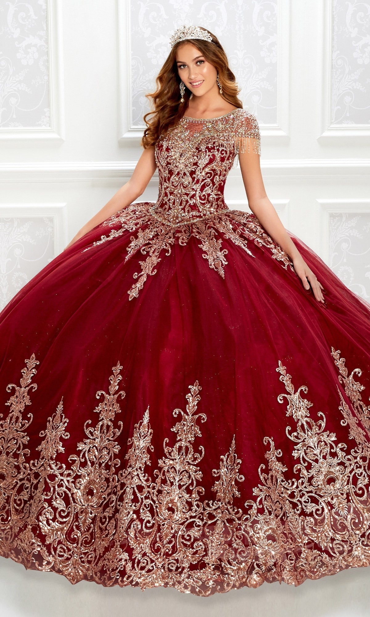 A woman wearing the Princesa PR22026 short-sleeve quinceanera dress with gold embroidery, a beaded bodice, and glitter tulle stands by a white ornate wall, smiling and accessorized with a matching tiara.